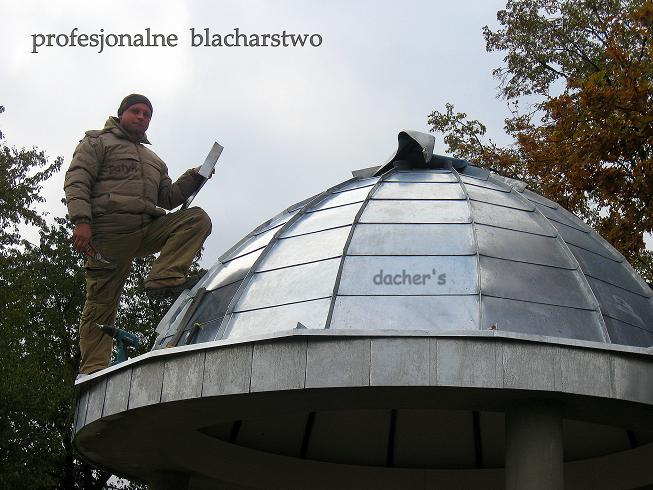 Pracownik w kurtce i spodniach roboczych, stojący na kopule z blachy, trzymający arkusz blachy, z wkrętarką i młotkiem przy pasie, podczas prac blacharsko-dekarskich.
